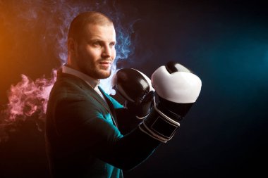 Genç yakışıklı işadamı adam beyaz gömlek, yeşil ceket, siyah ve beyaz boks eldiven gülümseyen ve kırmızı ve mavi siyah yalıtılmış zemin kağıttan duman karşı boks yapmak hazır