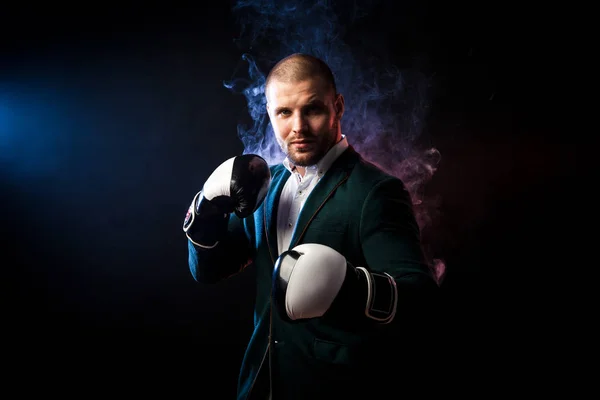 Young handsome businessman man in white shirt, green jacket, black and white boxing gloves smiling and ready to box against the red and blue smoke from the paper on a black isolated background