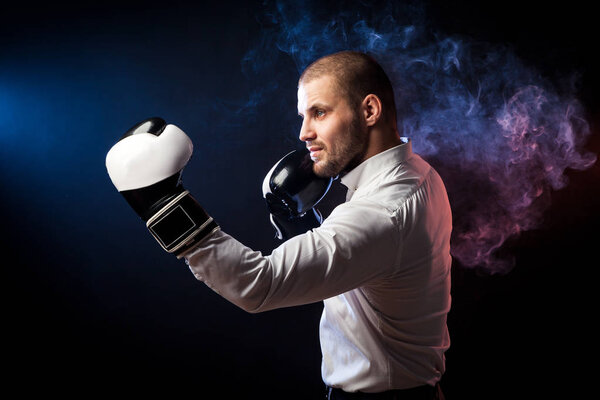 Красивый молодой человек бизнесмен в белой рубашке и черно-белых боксерских перчатках бокса перед красным и синим дымом от салфетки на черном изолированном фоне
