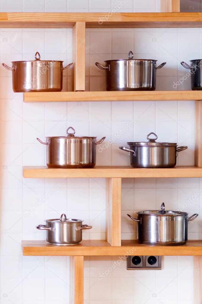 Beautiful modern mettalic new pans of different sizes stand on wooden shelves in the kitchen