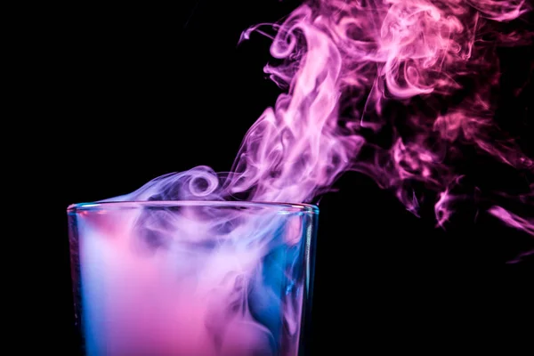 A close-up glass transparent glass filled with a wig from a blue, blue, purple wipe smokes and stands on a black isolated background. Glass bongs for smoking soft focus.