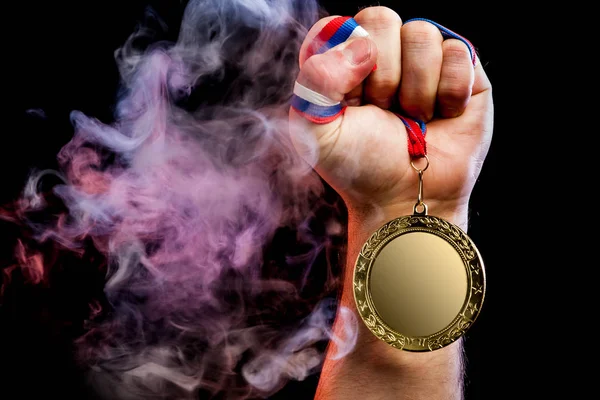 Close-up of a strong male hand holding a gold medal for a sporting achievement against a background of colored pink smoke and black isolated background. Mocap medals