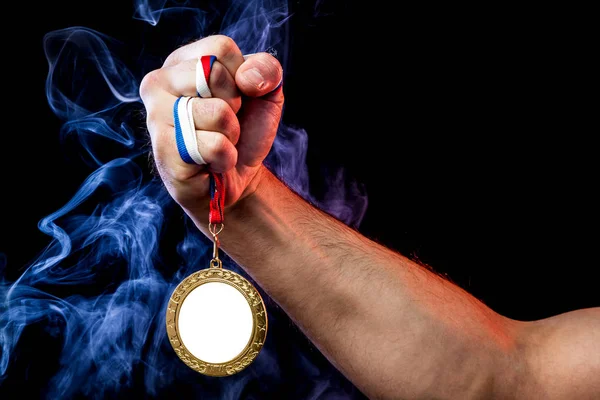 Close-up of a strong male hand holding a gold medal for a sporting achievement against a background of colored  blue smoke and black isolated background. Mocap medals