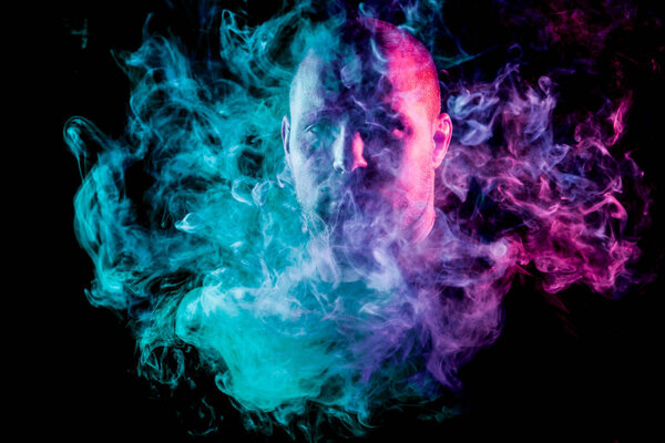 A young man exhales a cloud of colored smoke of blue and pink color on a black isolated. Harm to smoking on the example of a young mugger