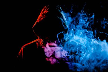 Siyah başlıklı genç bir adam sigara içiyor ve renkli yeşil ve pembe nefes vape izole bir siyah arka plan üzerine gelen duman
