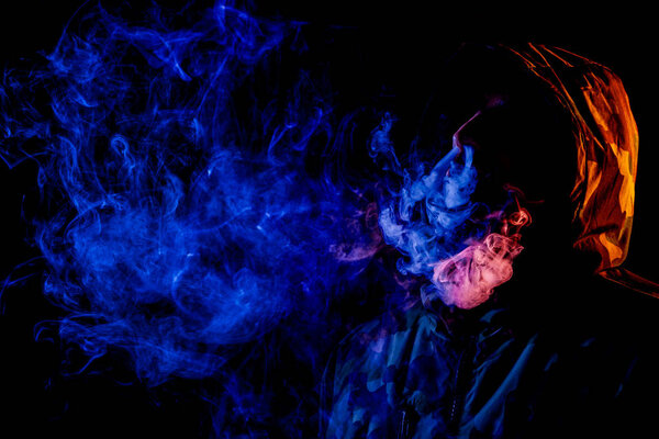 A young man in a black hood smokes and breathes out the colored blue and yellow  smoke from the vape a on a black isolated background