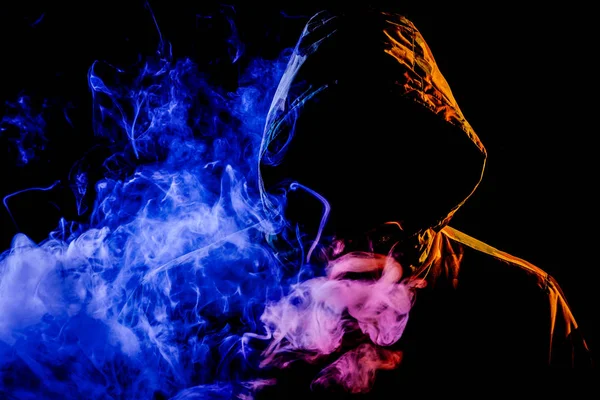 A young man in a black hood smokes and breathes out the colored  green  and pink smoke from the vape on a black isolated background