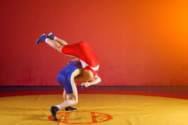 Kırmızı ve mavi üniformalı iki Yunan güreşçi, spor salonunda sarı bir güreş halısında güreşiyor.