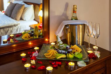 Sevenler için romantik akşam yemeği: bir meyve tabağı, bardak şampanya, şampanya buz metal bir kova ve mumlar, arka planda bir yatak ile bir tabloyla gül yaprakları dekore edilmiş