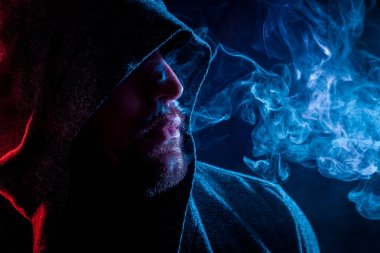 Siyah başlıklı yakışıklı bir adam sigara içiyor ve renkli mavi ve kırmızı siyah bir izole arka plan üzerinde duman büyük bulutlar exhales