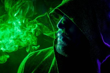 Siyah başlıklı yakışıklı bir adam sigara içiyor ve renkli yeşil ve mavi siyah bir izole arka plan üzerinde duman büyük bulutlar exhales