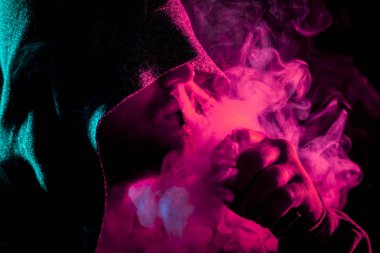 Siyah başlıklı yakışıklı bir adam sigara içiyor ve renkli pembe ve yeşil siyah bir izole arka plan üzerinde duman büyük bulutlar exhales
