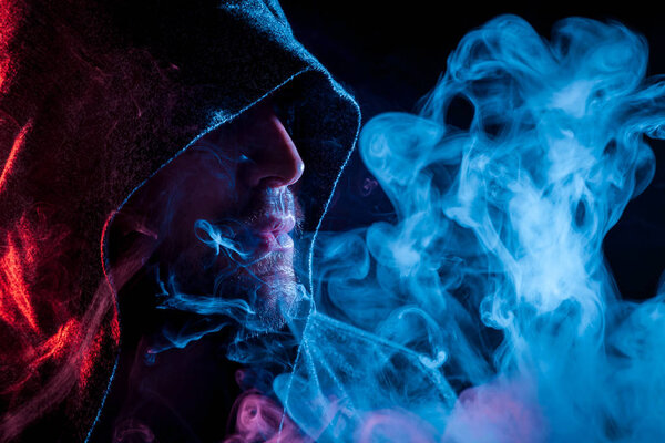 A young man in a black cape smokes and exhales large clouds of colored blue and red  smoke on a black isolated background