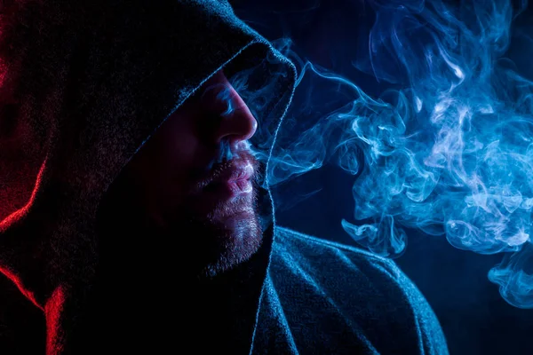 Siyah başlıklı yakışıklı bir adam sigara içiyor ve renkli mavi ve kırmızı siyah bir izole arka plan üzerinde duman büyük bulutlar exhales
