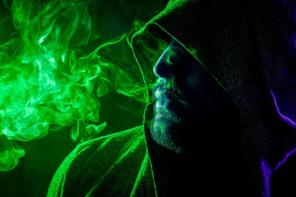Siyah başlıklı yakışıklı bir adam sigara içiyor ve renkli yeşil ve mavi siyah bir izole arka plan üzerinde duman büyük bulutlar exhales