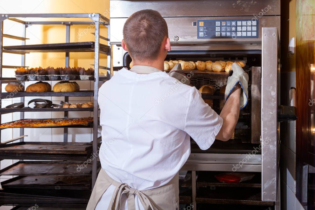 A male baker in white uniform and a beige apron bakes bread and takes cheese buns from an industrial oven. Work at the bakery