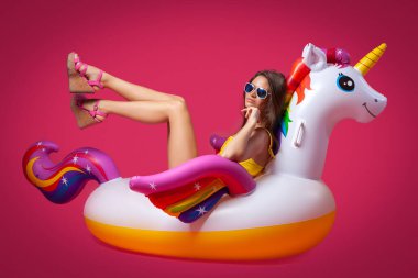 Yaz kıyafetleri içinde güzel zayıf bir kadın pembe izole edilmiş bir arka planda beyaz, şişme bir tek boynuzlu atta eğleniyor ve eğleniyor. Yaz havasını, rahatlığı ve güzelliği kavrayın..