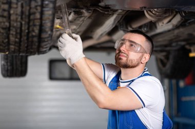 A young man auto mechanic in overalls at his workplace repairs the car's suspension, tightens the nuts on the chassis. Concept Worker at an automobile repair shop, a car lifted on a lift clipart