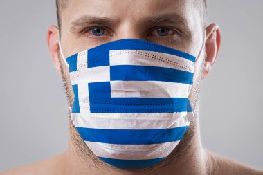Yunan bayrağının renklerine boyanmış tıbbi maskeli, yaralı gözlü genç bir adam.