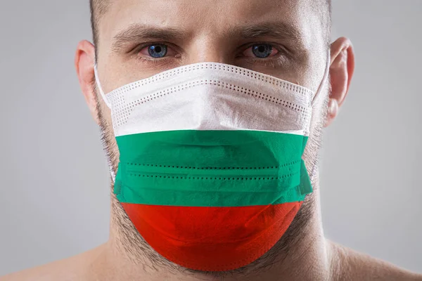 Young man with sore eyes in a medical mask painted in the colors of the national flag of Bulgari