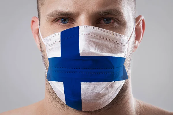 Young man with sore eyes in a medical mask painted in the colors of the national flag of Finlan