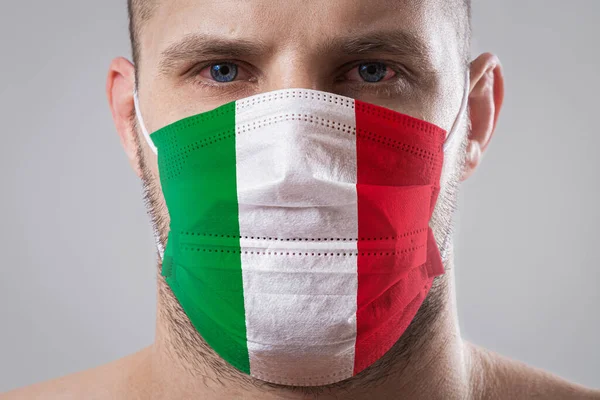 Young man with sore eyes in a medical mask painted in the colors of the national flag of Italy. Medical protection against airborne diseases, coronavirus. Man is afraid of getting the flu