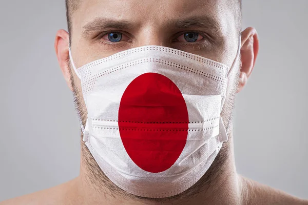Young man with sore eyes in a medical mask painted in the colors of the national flag of Japan. Medical protection against airborne diseases, coronavirus. Man is afraid of getting the flu