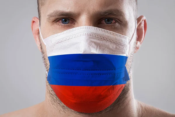 Young man with sore eyes in a medical mask painted in the colors of the national flag of Russia. Medical protection against airborne diseases, coronavirus. Man is afraid of getting the flu