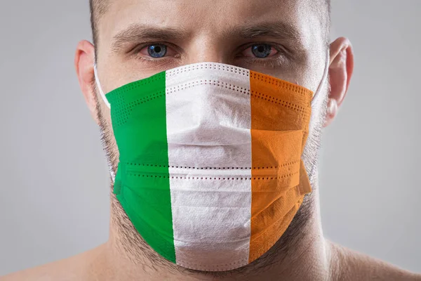 Young man with sore eyes in a medical mask painted in the colors of the national flag of Ireland. Medical protection against airborne diseases, coronavirus. Man is afraid of getting the flu