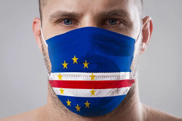 Young man with sore eyes in a medical mask painted in the colors of the national flag of Cape Verde. Medical protection against airborne diseases, coronavirus. Man is afraid of getting the flu