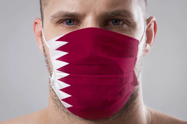Young man with sore eyes in a medical mask painted in the colors of the national flag of Qatar. Medical protection against airborne diseases, coronavirus. Man is afraid of getting the flu