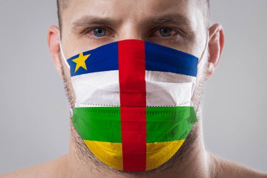 Orta Afrika Cumhuriyeti bayrağının renklerine boyanmış tıbbi maskeli yaralı gözleri olan genç bir adam. Hava hastalıklarına karşı tıbbi koruma, koronavirüs. Adam grip olmaktan korkuyor.