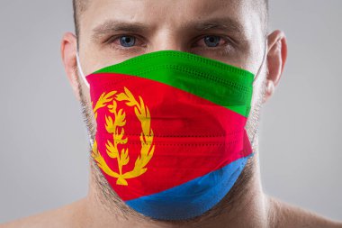 Eritre bayrağının renklerine boyanmış tıbbi maskeli, yaralı gözlü genç bir adam. Hava hastalıklarına karşı tıbbi koruma, koronavirüs. Adam grip olmaktan korkuyor.