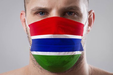 Gambiya bayrağının renklerine boyanmış tıbbi maske takmış yaralı gözleri olan genç bir adam. Hava hastalıklarına karşı tıbbi koruma, koronavirüs. Adam grip olmaktan korkuyor.