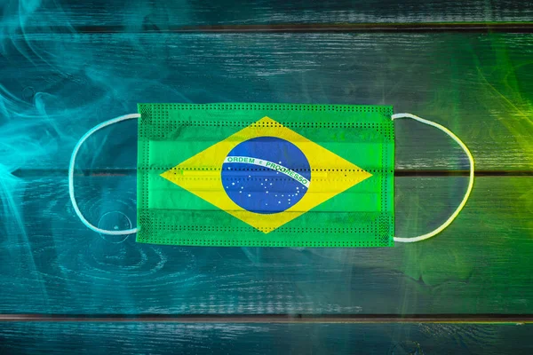 Medical mask for protection against airborne diseases, painted in the national flag of Brazil on a black background in blue-green smoke. Medical protection against airborne diseases, coronavirus