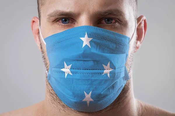 Young man with sore eyes in a medical mask painted in the colors of the national flag of Federal States of Micronesia. Medical protection against airborne diseases, coronavirus. Man is afraid of getting the flu