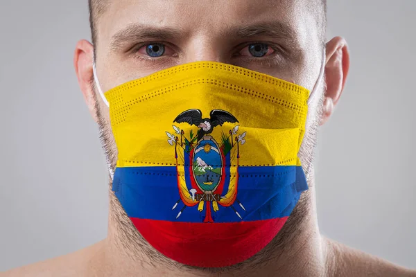 Young man with sore eyes in a medical mask painted in the colors of the national flag of Ecuador. Medical protection against airborne diseases, coronavirus. Man is afraid of getting the flu
