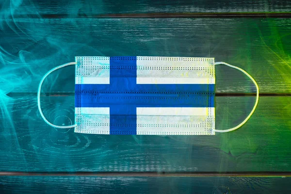 Medical mask for protection against airborne diseases, painted in the national flag of Finland on a black background in blue-green smoke. Medical protection against airborne diseases, coronavirus