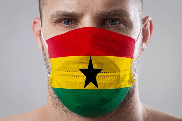 Young man with sore eyes in a medical mask painted in the colors of the national flag of Ghana. Medical protection against airborne diseases, coronavirus. Man is afraid of getting the flu