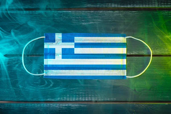 Medical mask for protection against airborne diseases, painted in the national flag of Greece on a black background in blue-green smoke. Medical protection against airborne diseases, coronavirus