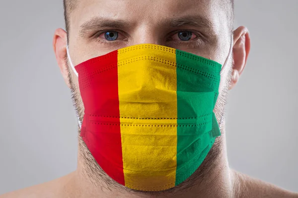 Young man with sore eyes in a medical mask painted in the colors of the national flag of Guinea. Medical protection against airborne diseases, coronavirus. Man is afraid of getting the flu