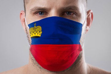 Lihtenştayn bayrağının renklerine boyanmış tıbbi maskeli, yaralı gözlü genç bir adam. Hava hastalıklarına karşı tıbbi koruma, koronavirüs. Adam grip olmaktan korkuyor.