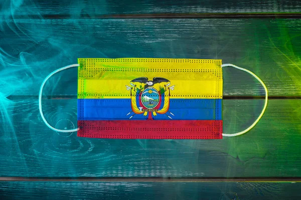 Medical mask for protection against airborne diseases, painted in the national flag of Ecuador on a black background in blue-green smoke. Medical protection against airborne diseases, coronavirus