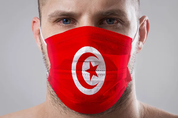 Young man with sore eyes in a medical mask painted in the colors of the national flag of Tunisia. Medical protection against airborne diseases, coronavirus. Man is afraid of getting the flu