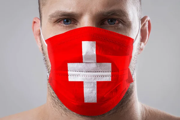 Young man with sore eyes in a medical mask painted in the colors of the national flag of Switzerland. Medical protection against airborne diseases, coronavirus. Man is afraid of getting the flu