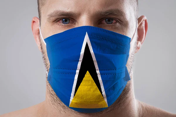 Young man with sore eyes in a medical mask painted in the colors of the national flag of Saint Lucia. Medical protection against airborne diseases, coronavirus. Man is afraid of getting the flu