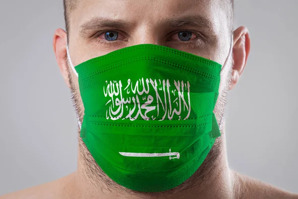 Young man with sore eyes in a medical mask painted in the colors of the national flag of Saudi Arabi