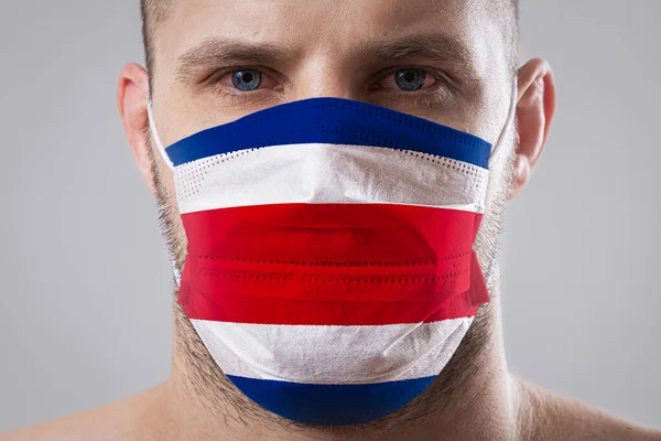 Young man with sore eyes in a medical mask painted in the colors of the national flag of Costa Rica. Medical protection against airborne diseases, coronavirus. Man is afraid of getting the flu