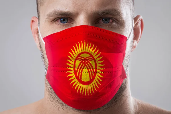 Young man with sore eyes in a medical mask painted in the colors of the national flag of Kyrgyzstan. Medical protection against airborne diseases, coronavirus. Man is afraid of getting the flu