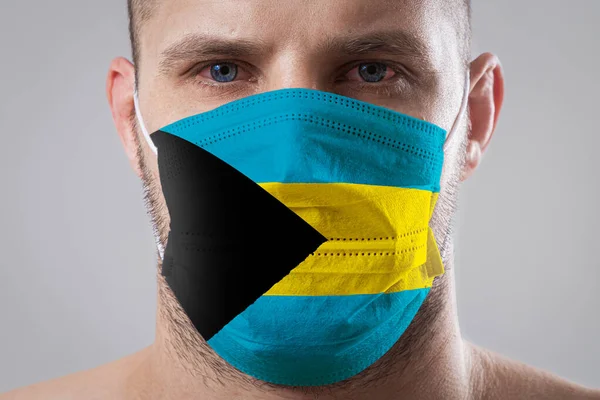 Young man with sore eyes in a medical mask painted in the colors of the national flag of Bahamas. Medical protection against airborne diseases, coronavirus. Man is afraid of getting the flu